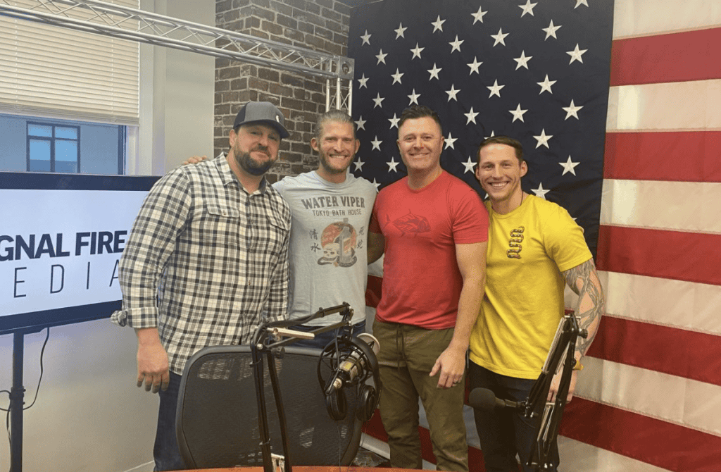 Diverse group of four men smiling in a recording studio with American flag backdrop and podcast equipment for Signal Fire Media.
