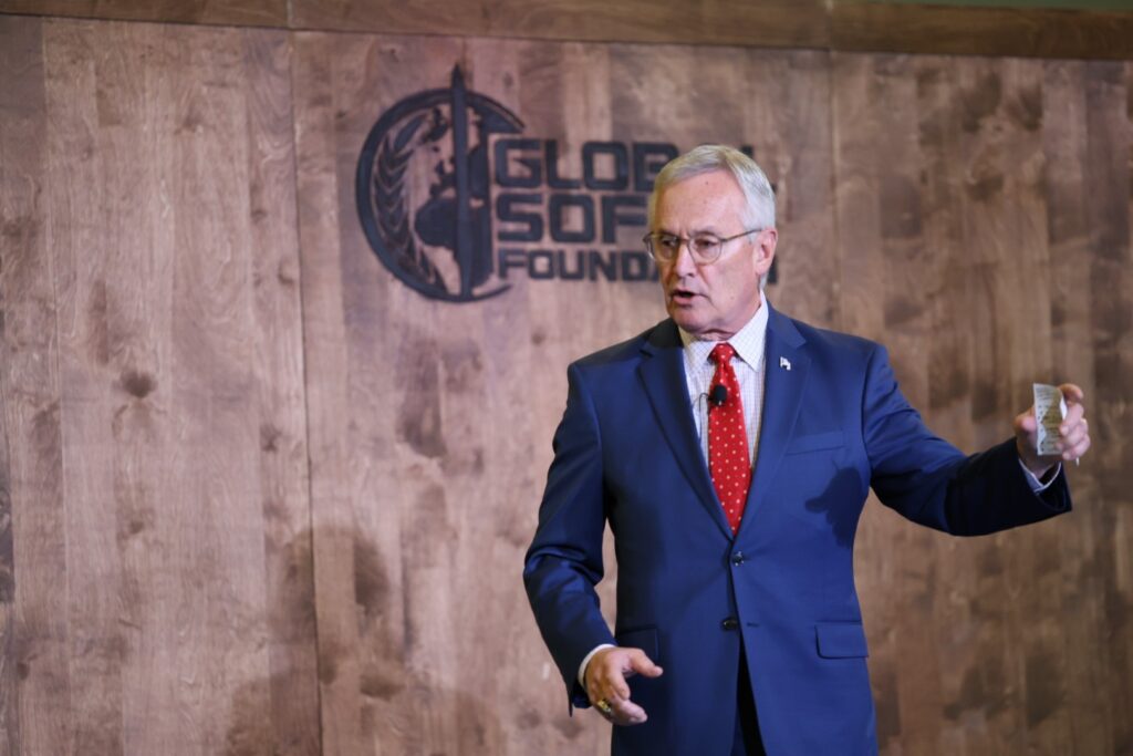 A man in a blue suit giving a presentation at the Global S.O.F. Foundation event, with a wooden wall and the foundation's logo visible in the background.
