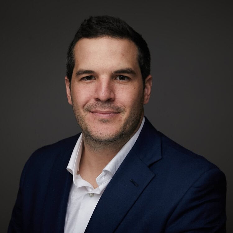 Professional headshot of a smiling man in a dark blue suit and white shirt, representing leadership in education and nonprofit sectors, shot against a dark gray background.