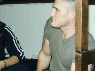 Man sitting under a bunk bed in an indoor setting, wearing a gray t-shirt and camouflage pants.