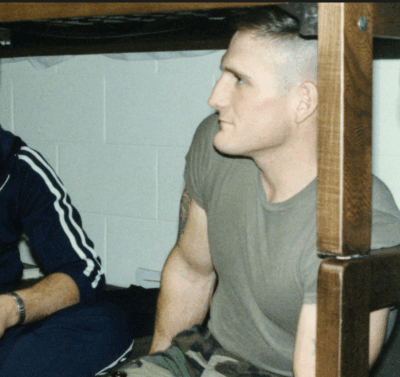 Man sitting under a bunk bed in an indoor setting, wearing a gray t-shirt and camouflage pants.
