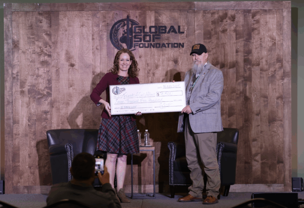 Check presentation of a large donation check at Global SOF Foundation event, featuring two individuals on stage, a woman with curly hair and a man with a beard and cap, in a professional setting.