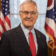 American man standing in front of American flags, wearing a suit and glasses, smiling for a formal portrait at GSOF event.