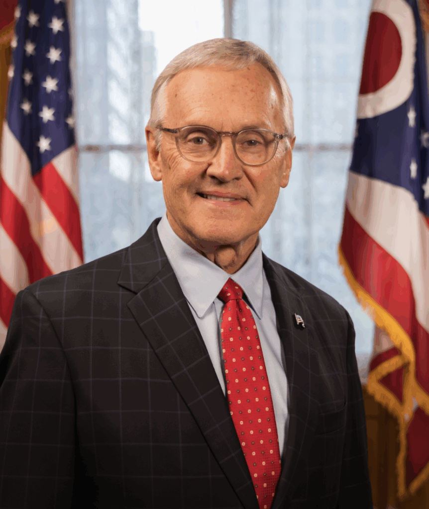 American man standing in front of American flags, wearing a suit and glasses, smiling for a formal portrait at GSOF event.